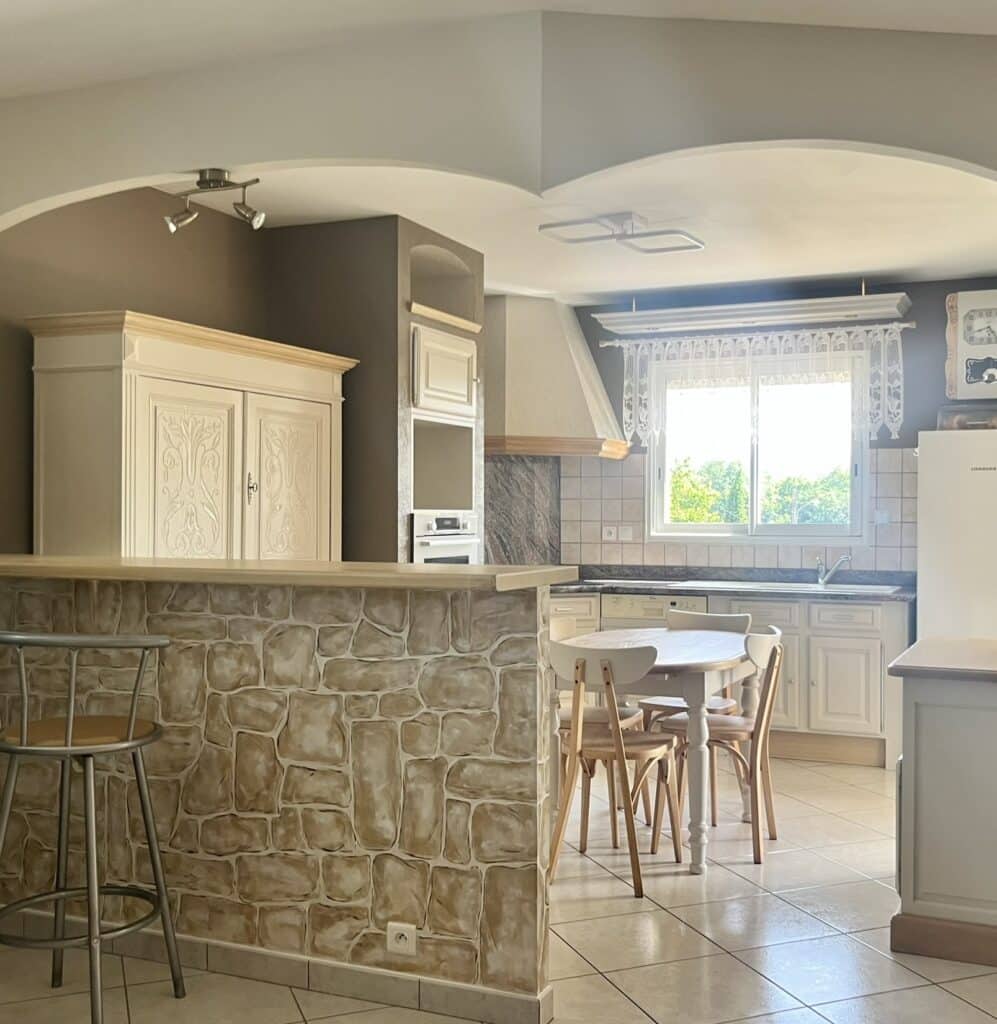 Cuisine rustique chic avec bar en parement pierre, table de repas blanche et grand buffet blanc sculpté.