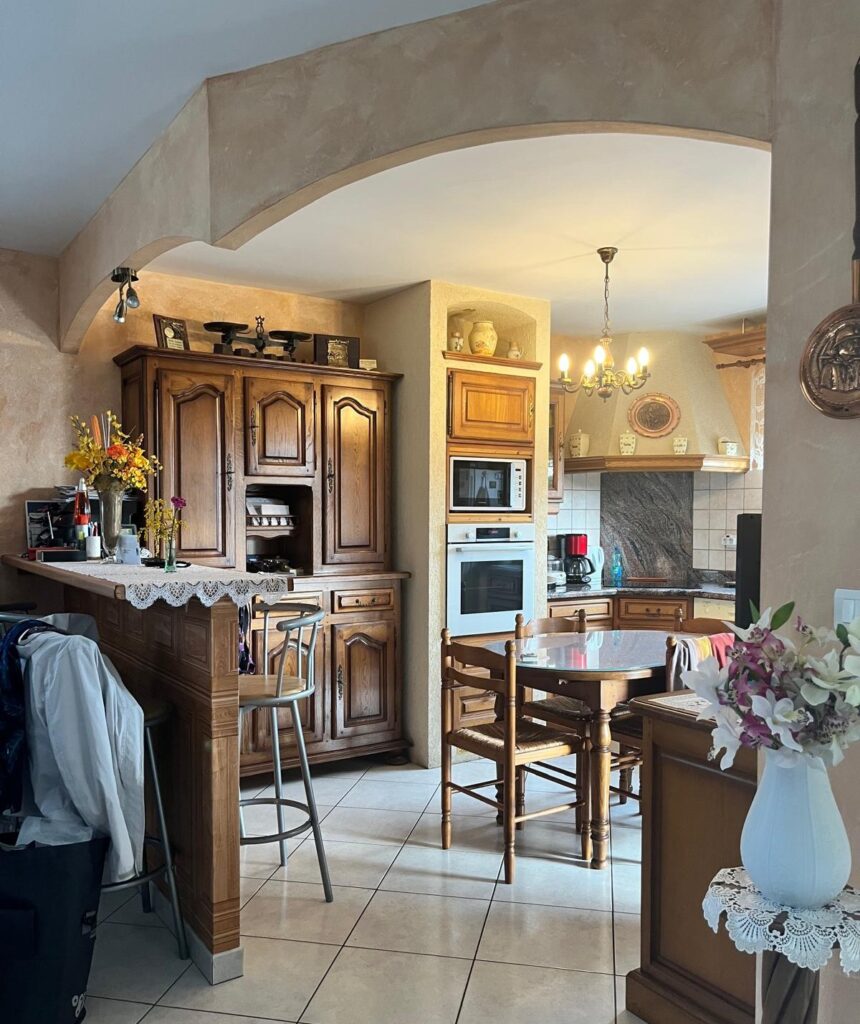 Cuisine rustique avec meubles en bois massif, table à manger, bar et arche décorative sous une lumière chaleureuse.