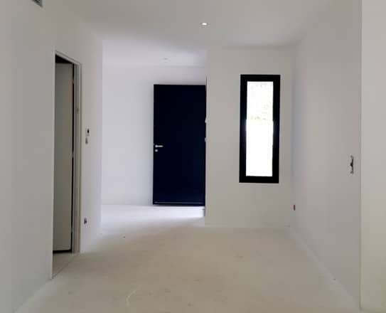 Intérieur blanc minimaliste en rénovation avec porte noire, fenêtre étroite et éclairage encastré.