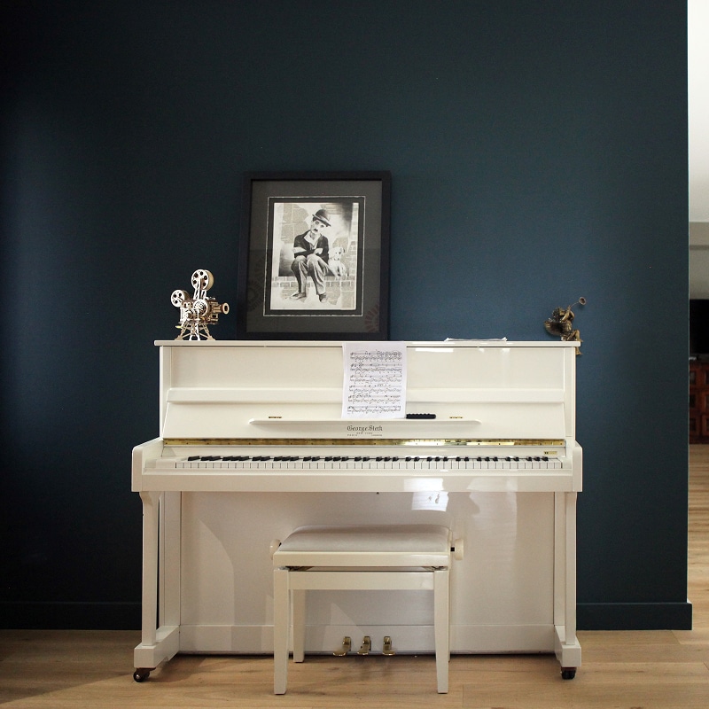 Piano George Steck Blanc sur Mur Bleu et Décoration Piano blanc George Steck devant un mur bleu foncé avec portrait de Charlie Chaplin et partitions de musique.