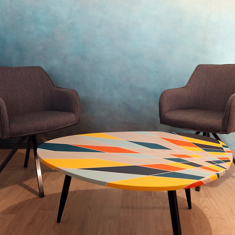 Table basse ronde design à motifs géométriques colorés Table basse ronde design à motifs géométriques colorés entre deux fauteuils gris sur fond de mur bleu texturé.